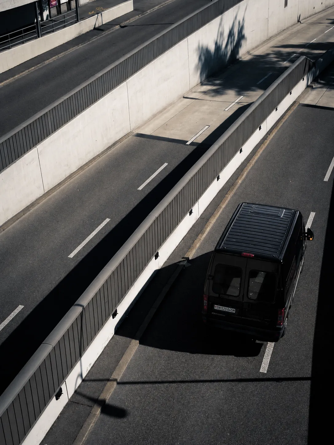 Black van driving in a sunlit urban divided road with shadows and concrete barriers.